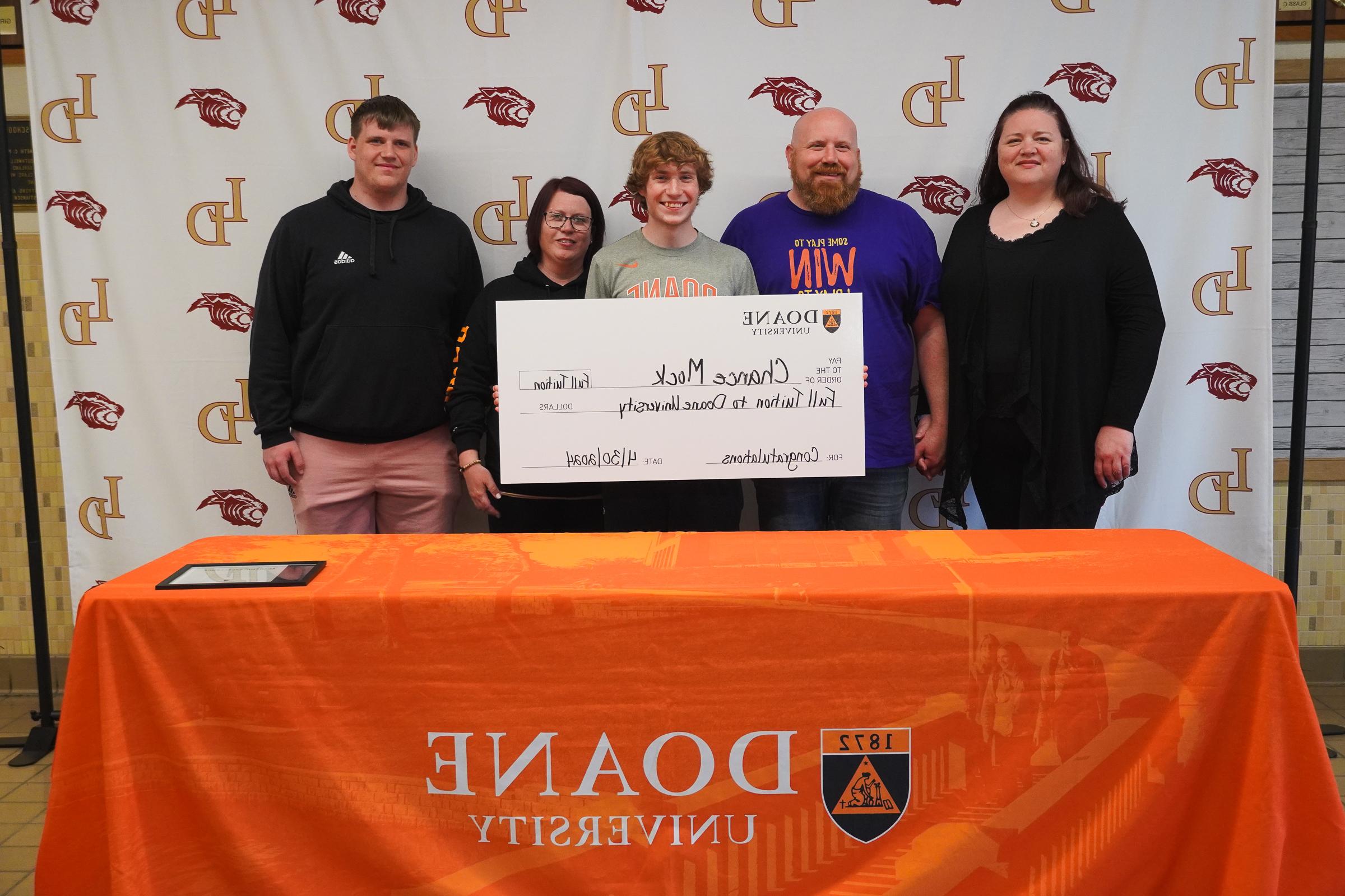 Five people stand in front of a white backdrop behind a table covered with an orange cloth. In the middle, a person holds up a giant check.