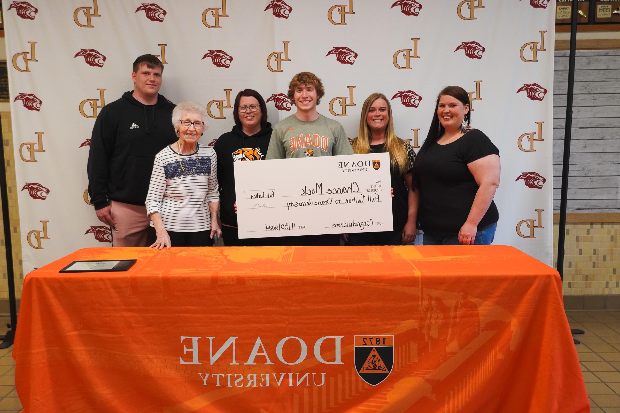 Six people stand behind a table covered in an orange cloth, in front of a white backdrop.