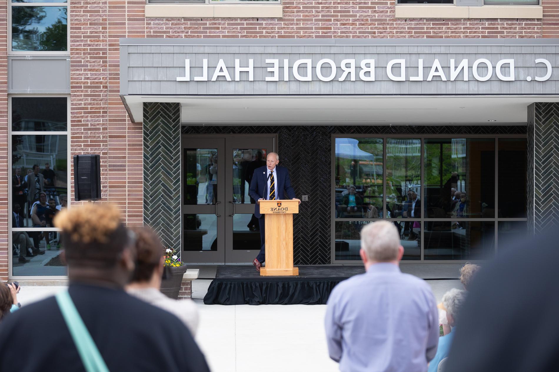 A man in a suit stands behind a wooden podium in front of an audience. Behind him is a building with a name reading "C. Donald 霍尔布罗迪."