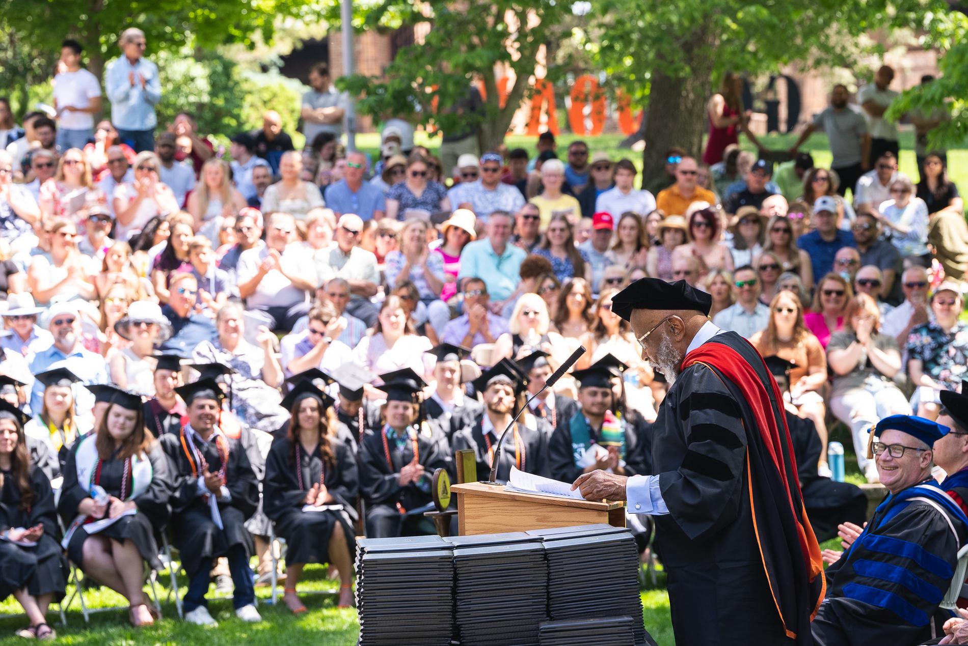 A Black man with a grey beard, wearing a black robe with a red 和 orange hood 和 a black cap, speaks to an audience from behind a podium. Members of the audience are smiling 和 clapping.