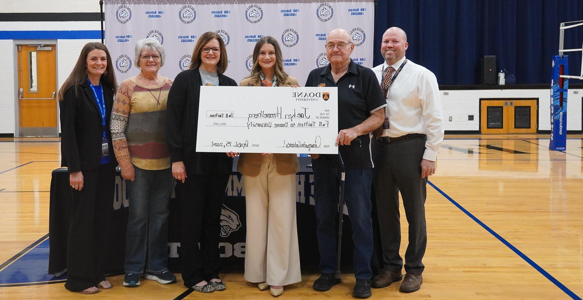 Six people stand in front of a white backdrop inside a school gymnasium. They're holding a large white board that looks like a check.